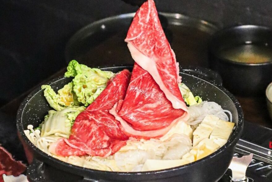 Thin slices of Wagyu beef being dropped into a pot of soup broth and vegetables cooking on a small cooktop.