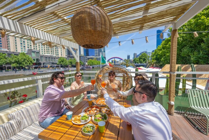 Group of people around a table on a venue floating upon the Yarra River, cheersing and eating food.