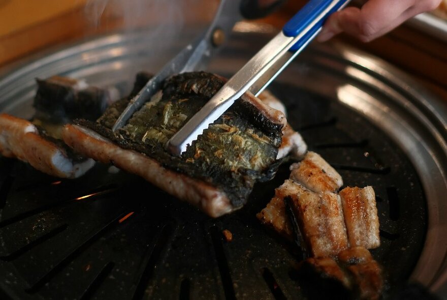 Hands holding a piece of grilled meat with tongs and using scissors to cut it into smaller pieces.