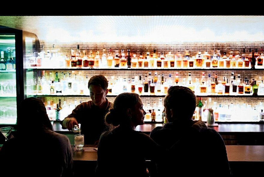 People seated at a backlit bar lined with shelves of bottles as a bartender pours drinks.