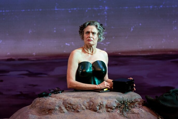 Performer Judith Lucy from the waist up, her lower half buried in a small hill, on a stage.