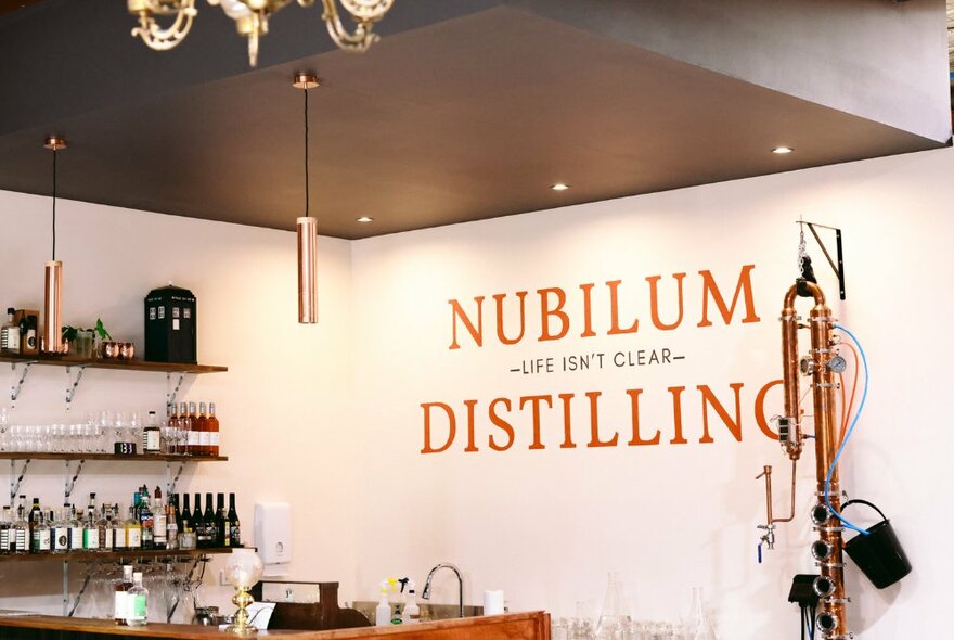 A small bar with shelves of alcohol and drinking equipment, and a painted sign on the back wall that says NUBILUM DISTILLING.