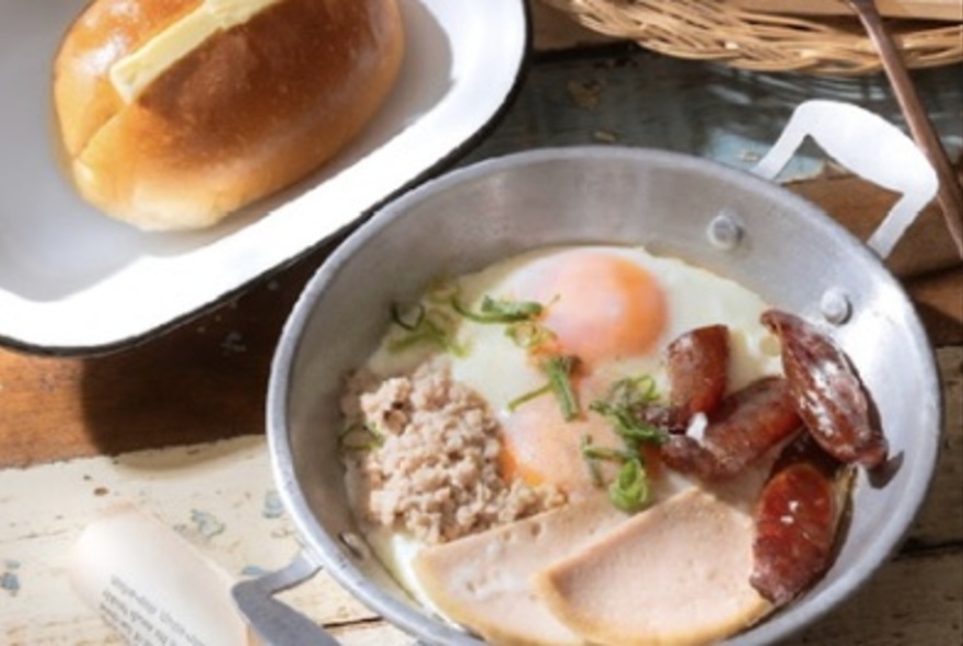 A cooking dish of egg and meat next to a buttered bun in a dish.