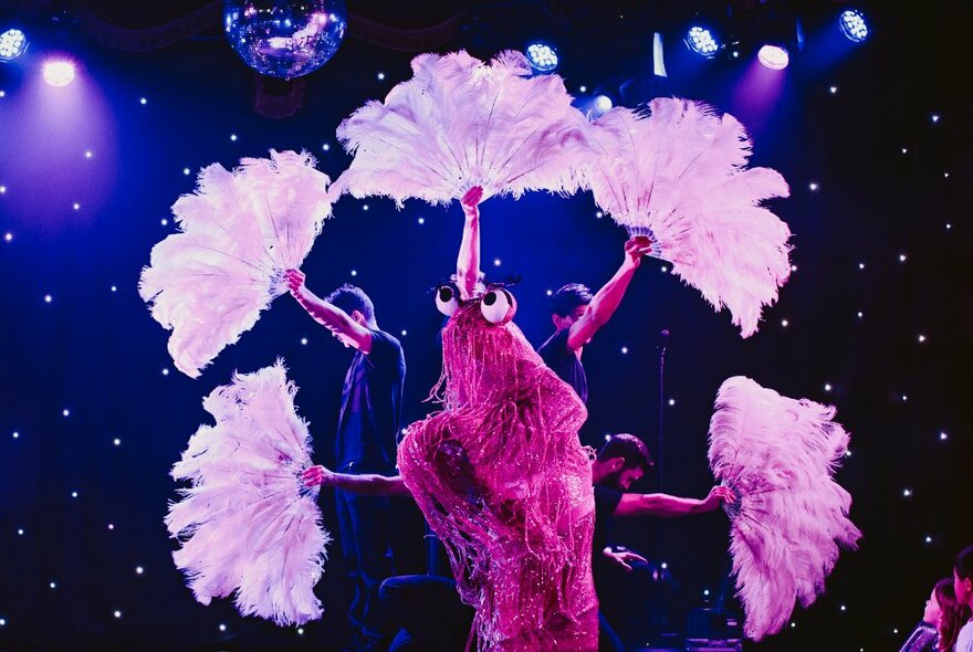 Performers on a darkened, starry stage holding out pink feather fans in a circular shape.