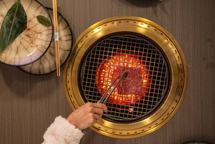 Hands holding tongs and cooking marbled beef on a gold-rimmed Japanese tabletop barbecue.