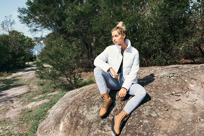 Woman in parka, jeans and lace up boots seated outdoors on a rock.