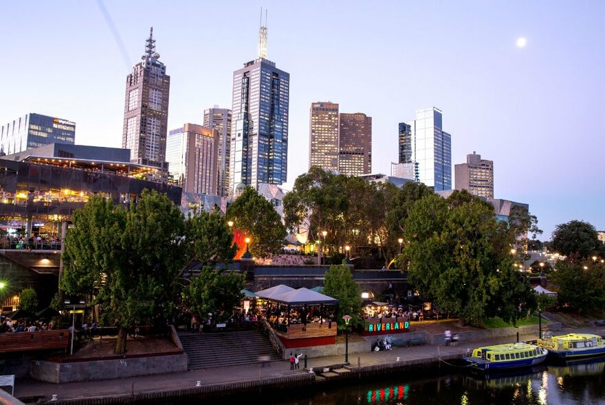 Melbourne city buildings behind trees and restaurants lining the Yarra River.