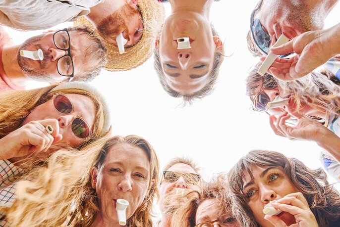 View taken from below and looking up at a group of people with their heads bent close together in a loose circle, and all blowing into kazoos that they hold in their mouths.