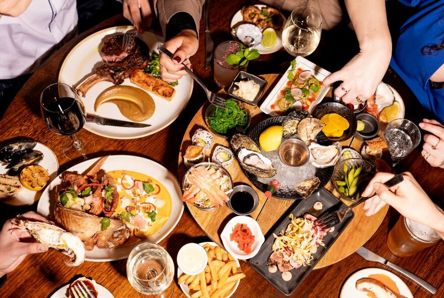 An overhead view of a table filled with plates of food and drink with people's hands using cutlery to select food. 