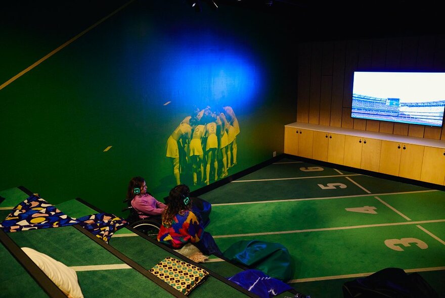 Two women sitting on indoor tiered seating decorated to look like a sports field, watching a screen with headphones on. 