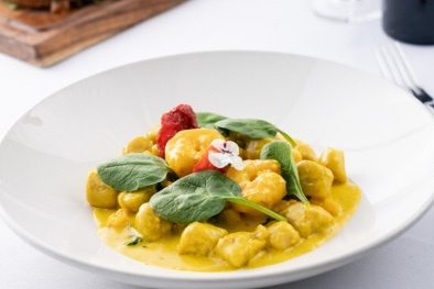White shallow bowl of gnocchi with creamy yellow sauce, garnished with several baby spinach leaves and edible flowers.
