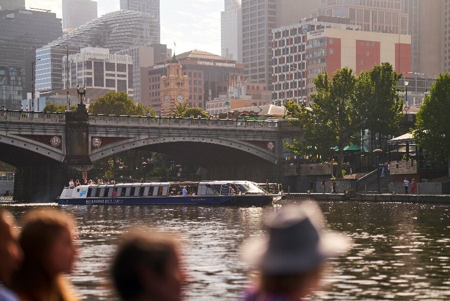 People standing on a riverside watching a riverboat passing under a bridge, with city buildings in the background.
