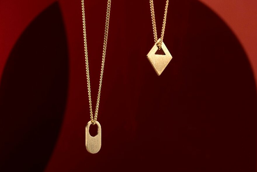 Two small pendants hanging off gold chains against a dark red-brown background.