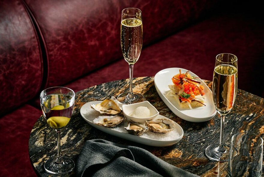 Two champagne glasses, one sherry glass and two obong plates with food, on dark marble-look surface.