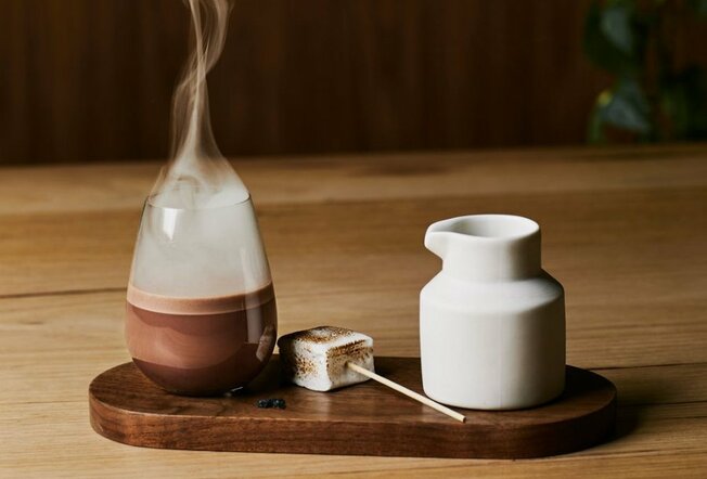 A hot chocolate in a glass with steam coming off it, next to a marshmallow and milk jug.