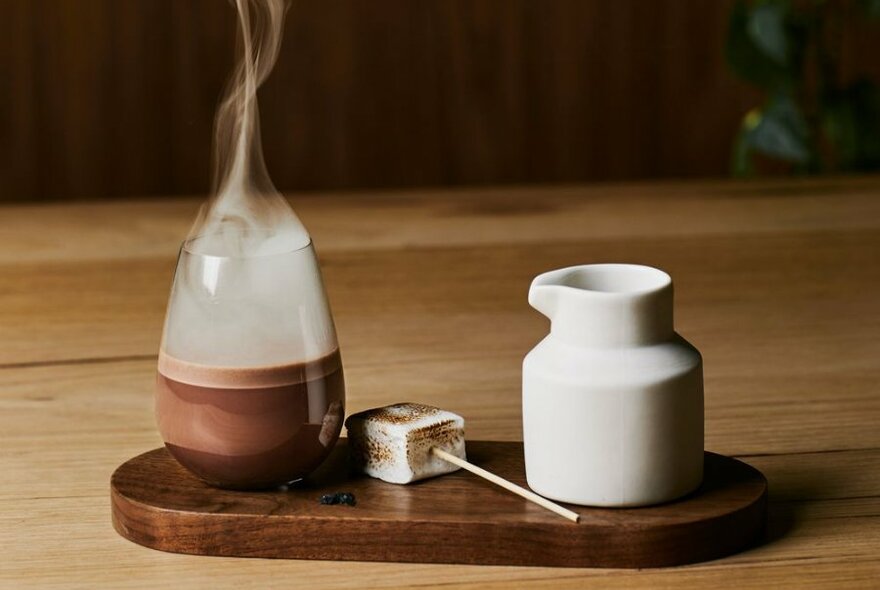 A hot chocolate in a glass with steam coming off it, next to a marshmallow and milk jug. 