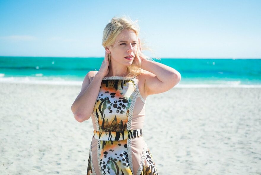 Woman wearing a patterned sun-frock, standing on a sandy beach, turquoise coloured sea in the background, holding her blond hair down with her hands.