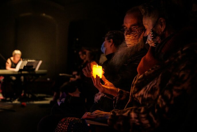 Two people in foreground holding a candle in dark room, other audience members and person at back on piano, blurred.