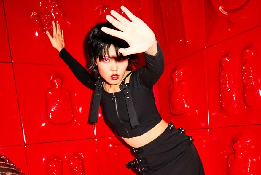 A model posing in front of a 3d red plastic wall, wearing black clothing covered in straps