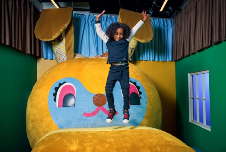 A young child jumping on a giant, plush, yellow bunny with a blue face in a small green room with a window.