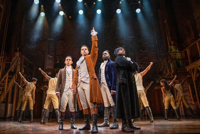 The cast of Hamilton on stage under spotlights, gesticulating and wearing 18th-century costumes.