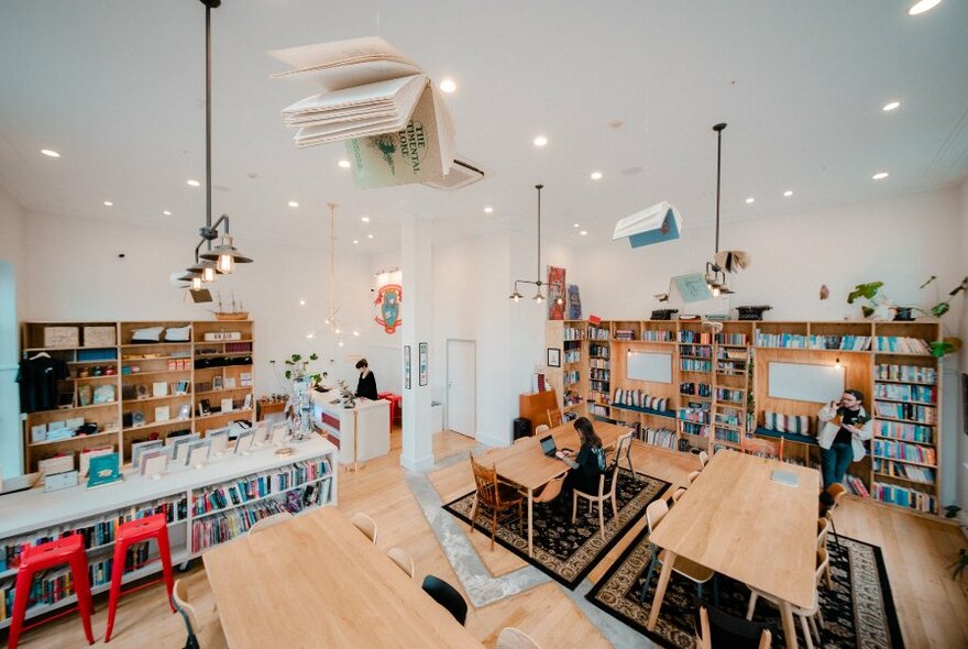 Birds eye view of a bright and airy writing studio space, with walls of bookshelves, white walls, large tables and writing benches, chairs, rugs and people working on laptops.