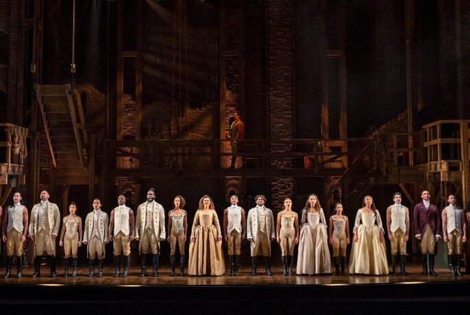 The cast of Hamilton standing in a row on stage with arms by their sides, three women wearing crinolines standing at the front.