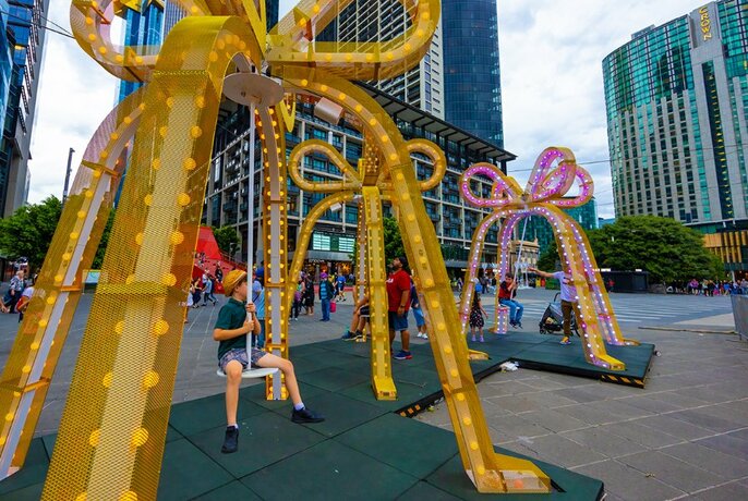 Children playing on giant swings shaped like bells with city buildings in the background.