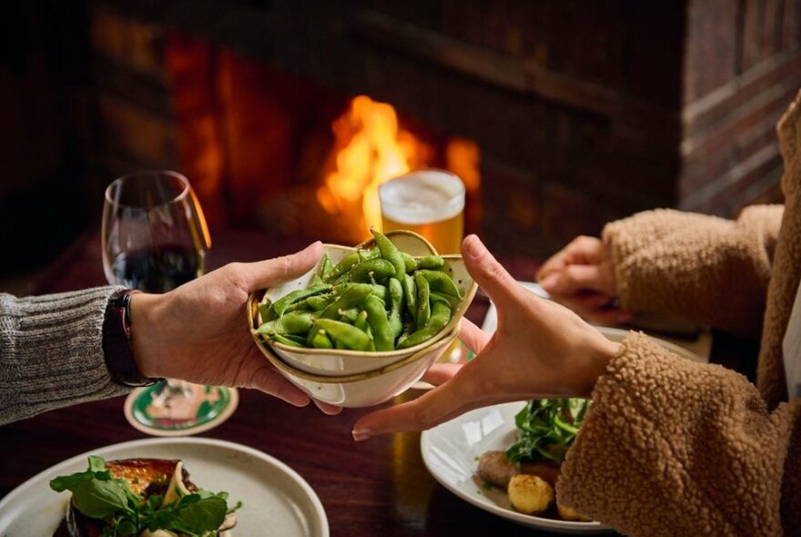 A couple handing each other a plate of food in front of a fireplace at a restaurant