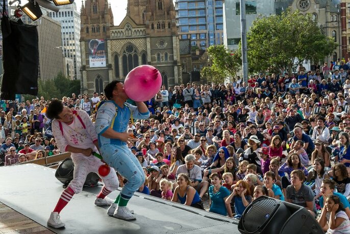 Clowns performing on a stage in Fed Square, blowing up balloons in front of a huge crowd.