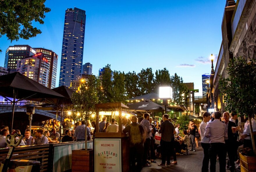 Exterior of Riverland Bar showing seated and standing patrons at outdoor tables with a background view of city skyline at dusk.