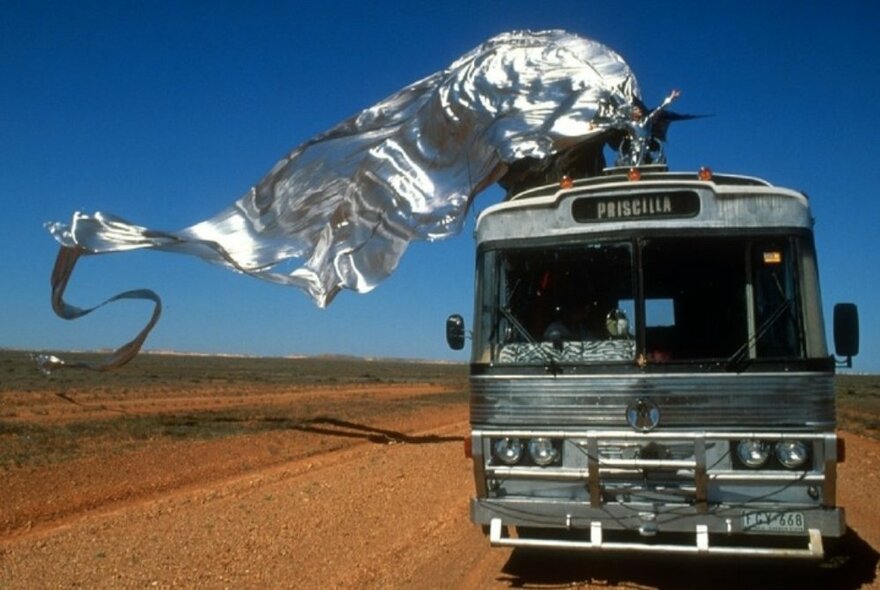 The big silver bus from the film The Adventures of Priscilla, Queen of the Desert, driving along a long red empty road in the outback, with a person standing on the roof of the bus wearing a flowing silver cape that billows in the wind behind the bus.