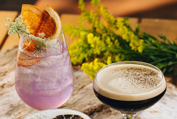 An espresso martini and a lavender coloured cocktail with dried citrus garnish with a bunch of wattle in the background