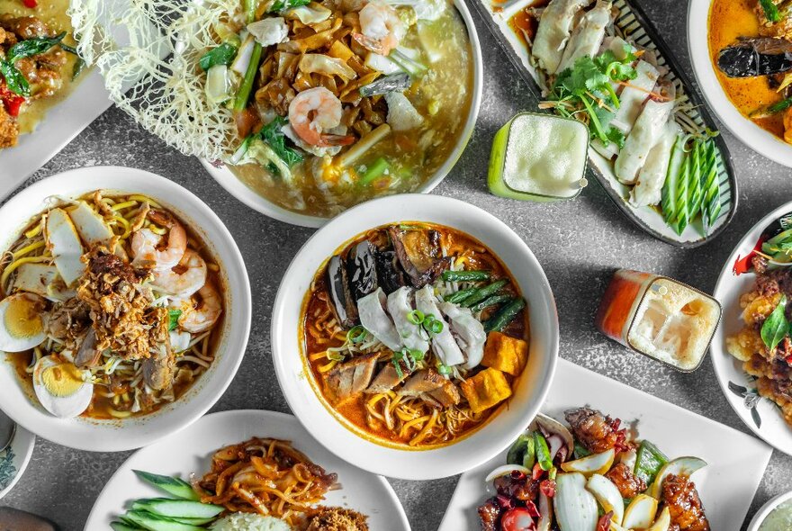 An overhead view of a table laden with an assortment of Asian dishes of food including seafood and egg noodles, salads, soups and dips.