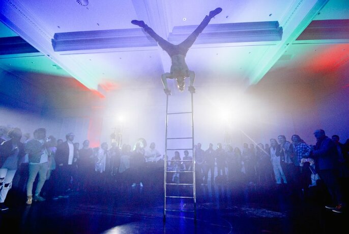 Audience standing in semi-circle around person upside down atop a ladder, in bright white light.