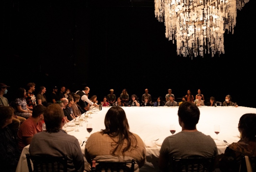 A stage-sized table with people seated all around it in chairs, glasses of wine and cutlery in front of some of the audience.
