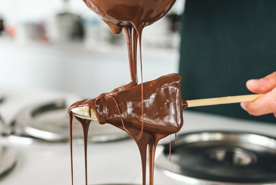 Melted chocolate being poured over a wedge of cheesecake on a stick. 