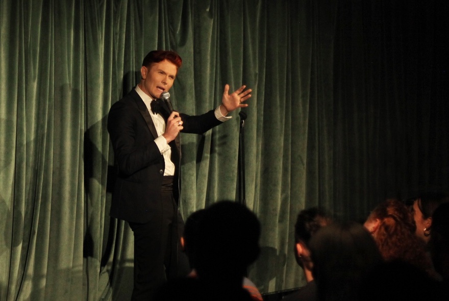 Comedian Rhys Nicholson on a stage wearing a black suit and white shirt, talking into a microphone the yhold, audience watching.