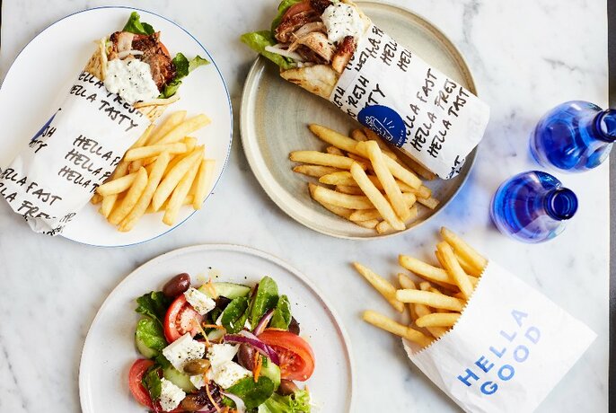 Souvlakis and chips wrapped in paper, a Greek salad and blue bottles on a marble table