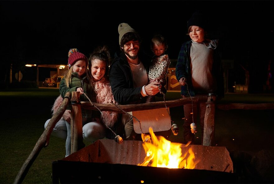 A family dressed in warm winter gear roasting marshallows over an open fire.