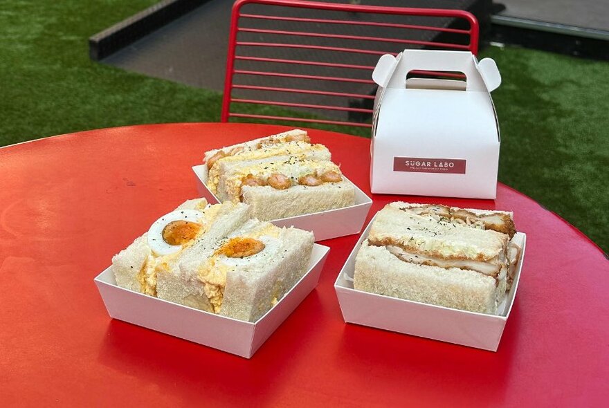 Japanese filled sandwiches or sandos in white cardboard takeaway containers on a red table.