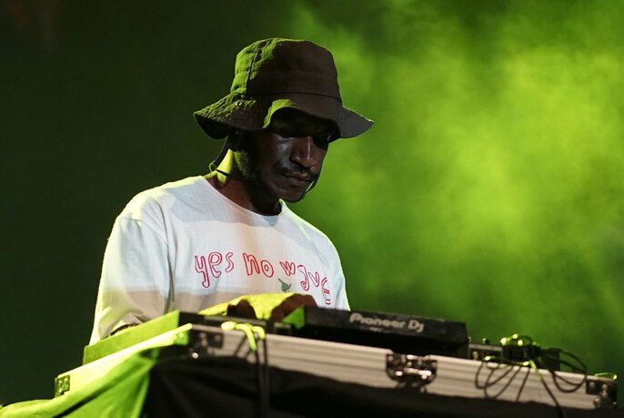 Man wearing a dark green hat pulled down low over his head, looking down and operating a DJ mixing desk with ambient green-yellow smoky haze behind him.