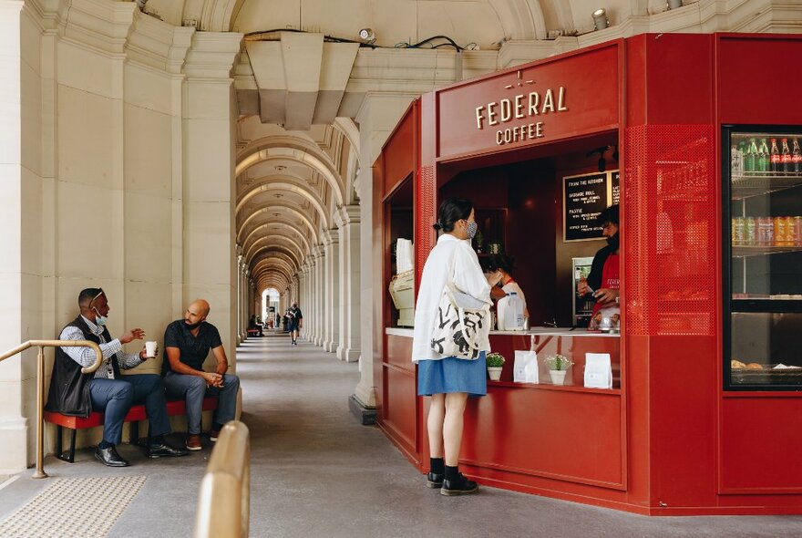 The archways of the Melbourne GPO Building with a red shopfront and a customer ordering coffee, two men seated on a red bench to the left.