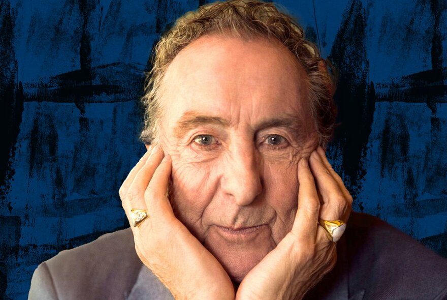 Comedian Eric Idle, staring directly at the viewer with a wry smile, his head resting in his hands, which are cupped around his chin and cheeks.