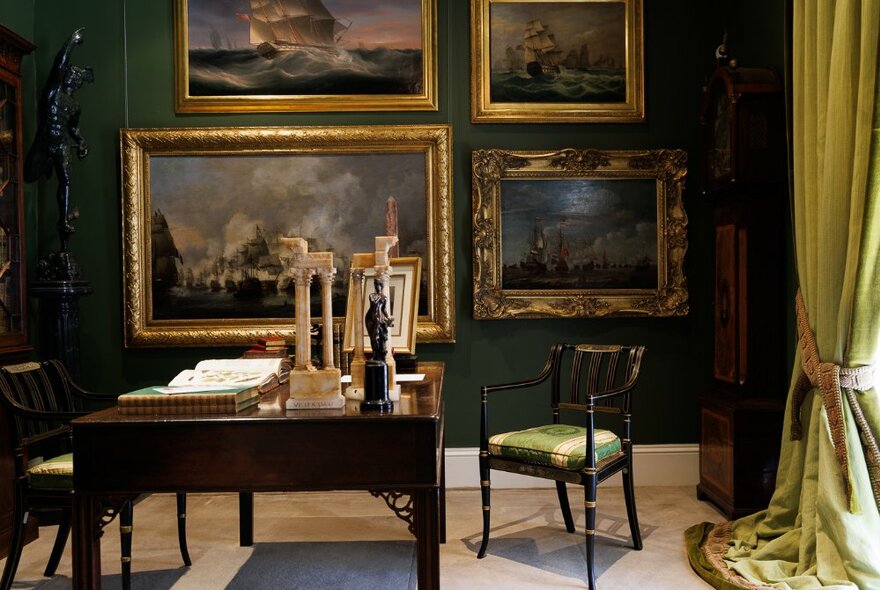 A dark Georgian drawing room with a desk, a chair pulled out  and large gold frames on the walls