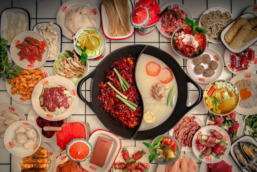Overhead view of a table laden with many small plates of fresh and raw ingredients, waiting to be cooked in a hot pot in the centre of the table.