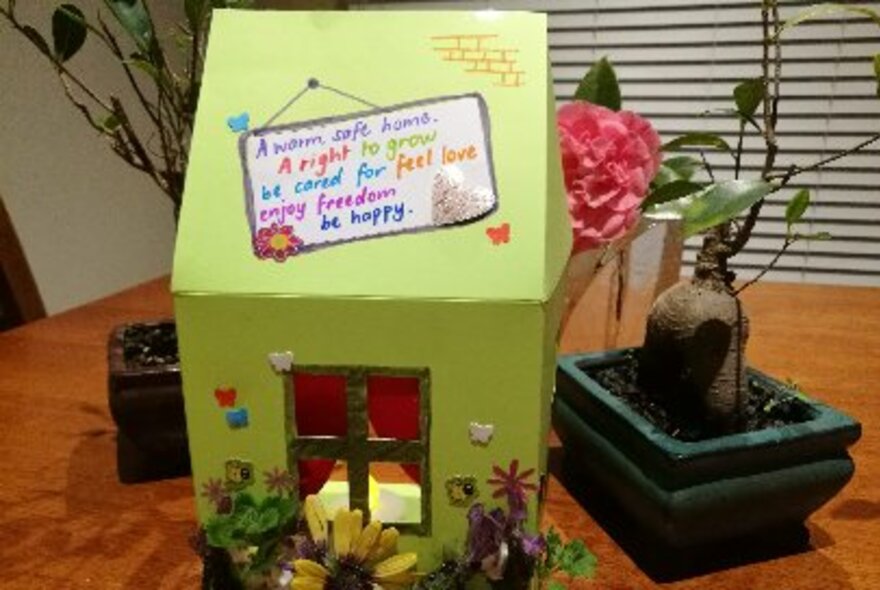 A colourful, hand decorated cardboard home with paper flowers and a handwritten message on the roof. 