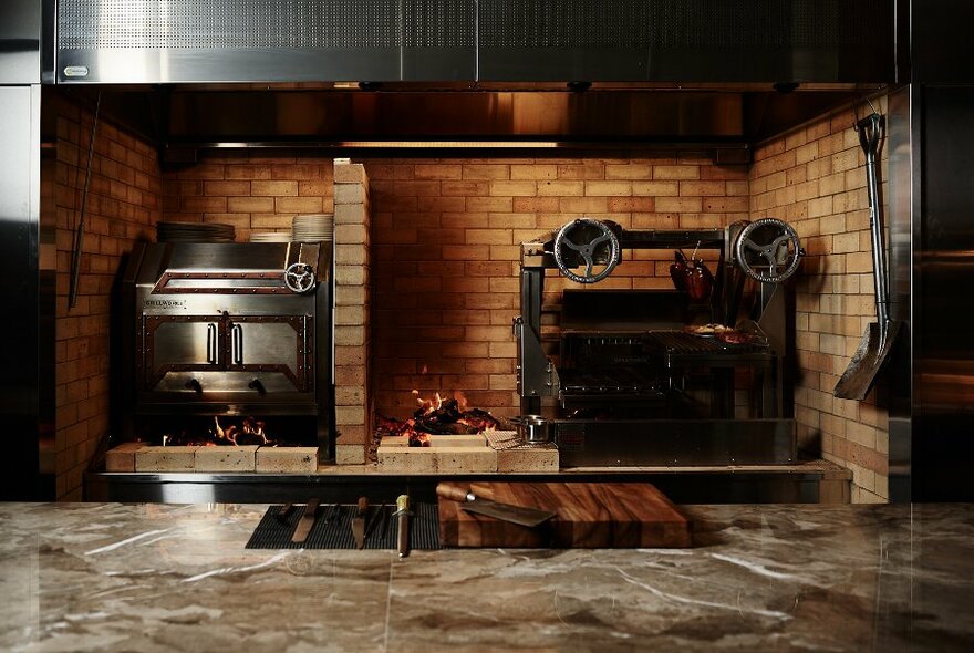 Large industrial metal oven, next to small open fire, next to metal industrial contraption, seen over marble bench.