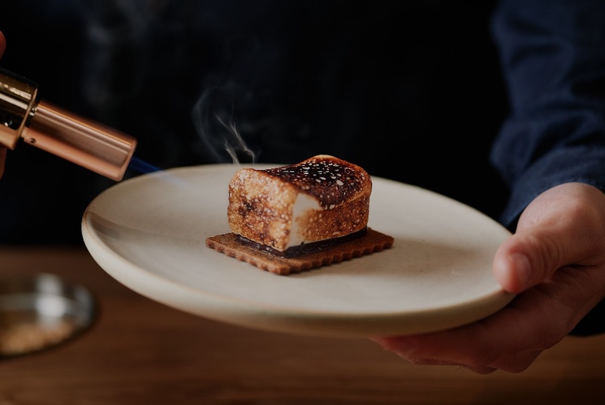 Small blow torch held to left of chocolatey-marshmallowy dessert, on white plate held by hand, blue-clothed arm and torso behind.
