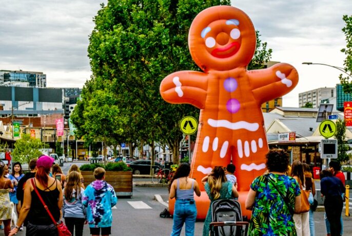 Giant orange gingerbread inflatable, in an open plaza area of a market with people walking around, a large green tree in the background.
